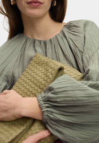 Pochette intrecciata verde oliva tenuta da una persona, abbinata a una blusa leggera con texture che presenta maniche a sbuffo e un colletto arricciato.