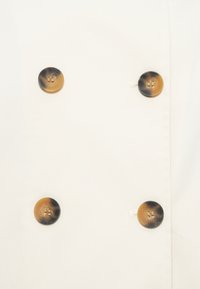 Tissu blanc à texture lisse présentant quatre gros boutons ronds brun foncé avec des centres plus clairs, espacés également sur l'avant.