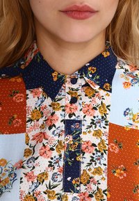 Geknoopte blouse met een patchworkdesign in bloemmotieven en stippen. Kleuren zijn marineblauw, oranje, lichtblauw en wit.