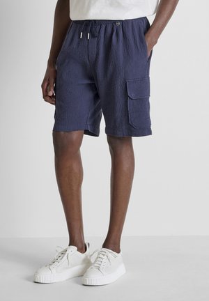 Hombre con pantalones cortos tipo cargo texturizados de color azul marino con cintura elástica y zapatillas blancas, de pie frente a un fondo liso y claro.