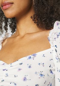 Blusa branca floral com pequenos padrões de flores azuis e roxas, apresentando mangas bufantes e um design com pregas na frente. Textura suave.