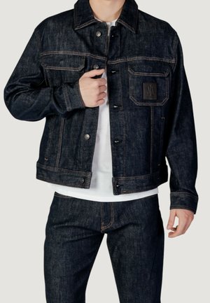 Mann trägt dunkelblaue Jeansjacke und passende Jeans über einem weißen Hemd, richtet mit der linken Hand den Jackenkragen.