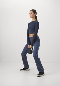 Tengerészkék, hosszú ujjú crop top, amelyhez illeszkedő, magas derekú nadrágot visel a modell. A modell fekete kettlebellt tart a kezében, fekete sportcipőt visel.
