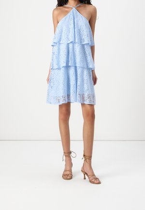 Kobieta w jasnoniebieskiej koronkowej sukience na ramiączkach z falbanami, z beżowymi sandałami na obcasie z paskami, na białym tle.