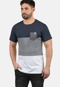 Camiseta de cuello redondo azul marino con rayas horizontales en el centro, un bolsillo gris y una sección inferior blanca. Tejido de mezcla de algodón suave.