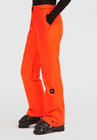 Persoon in feloranje sneeuwbroek met zwarte skibotten en een zwart langarmshirt, met de handen in de zakken, staand op een witte ondergrond.
