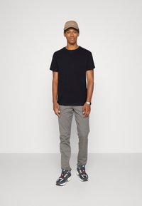Svart t-skjorte, grå bukser og flerfargede sneakers. Lys brun caps og klokke som bemerkelsesverdige tilbehør. Nøytral bakgrunn.