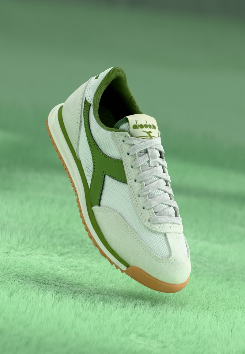 White and green sneaker with mesh and suede materials, round toe, lace-up design, and orange rubber sole, featuring a logo on the tongue.