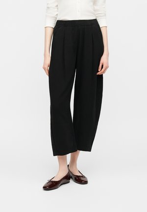 Žena nosí voľné čierne nohavice po členky, bielu košeľu s dlhým rukávom a tmavohnedé lakované balerínky.