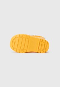 Semelle de chaussure en caoutchouc jaune présentant une surface texturée avec un motif de semelle et des détails de marque en relief. Bords lisses et forme arrondie.