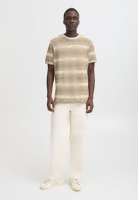 Stripete crew-neck t-skjorte i beige og dempet gul, med horisontale linjer. Kombinert med beige, løstsittende bukser og lyse joggesko.
