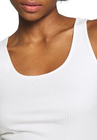 Vitt ribbat linne med bred halsringning och axelremmar, med en slät textur och en kroppsnära passform som framhäver hudtoner.