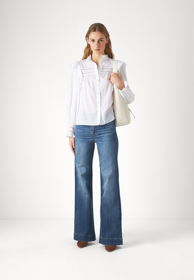 Vit skjorta med spetsdetaljer, ihopparad med utsvängda blå jeans och bruna skor. Modellen håller en krämfärgad handväska. Enkel bakgrund.