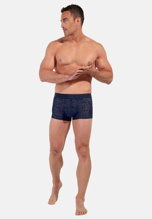 Mørkeblå bokserbriefs med et blonderlignende mønster, blød tekstur og en bred, mærkevarebælte. Modellen poserer med et neutralt ansigtsudtryk.