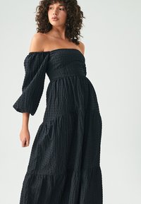 Svart off-the-shoulder klänning med texturerad tyg, lager-på-lager kjol och trumpetärmar. Designen har ett figursytt liv och en flödande silhuett.