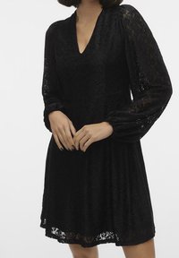 Svart spetsklänning med långa puffärmar, V-ringning och A-linjeformad silhuett, med ett texturerat blommönster. Sl ät tygfinish.