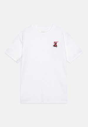 Balta kokvilnas T-krekls ar mazu izšūtu lāci un vārdu "Guess" sarkanā krāsā virs tā. Īsās piedurknes un apaļa kakla izgriezums.