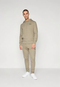 Sweatshirt bege com capuz, bolso canguru e cordão, combinado com calças de jogging justas a condizer. Apresenta o logótipo "BORG" em preto, complementado por ténis brancos.