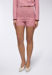 Pantaloni corti rosa su misura con una texture liscia, vita alta e tasche laterali, abbinati a tacchi bianchi lucidi. Presentano un design aderente.