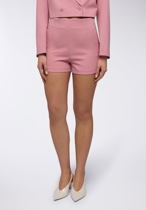 Pantaloni corti rosa su misura con una texture liscia, vita alta e tasche laterali, abbinati a tacchi bianchi lucidi. Presentano un design aderente.