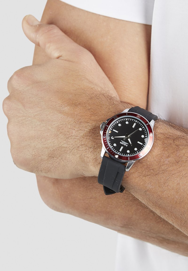 Montre noire avec bracelet en caoutchouc, accents en acier inoxydable et cadran rond noir comportant des marqueurs d'heure blancs et une lunette rouge.