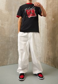 Černé bavlněné tričko s červenými akcenty a grafickým potiskem. Bílé cargo kalhoty s kapsami. Černé a červené tenisky s bílou podrážkou.