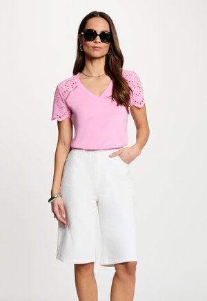 Kvinde iført en lyserød kortærmet top med hulbroderidetaljer, hvide knælange shorts, sorte solbriller og sølvsmykker, stående med hånden i lommen.