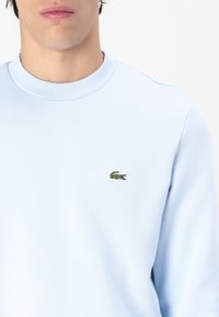 Sweat-shirt bleu clair à col rond en tissu doux, avec un petit logo crocodile vert brodé sur la poitrine gauche.