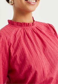 Blouse rouge avec un col à volants, tissu texturé présentant des lignes horizontales et une broderie florale, manches courtes et coupe décontractée.