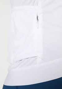 Top atlético blanco con un bolsillo lateral con cremallera, textura suave y un diseño ajustado. El dobladillo es ligeramente curvado para facilitar el movimiento.