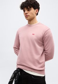 Νεαρός άνδρας με κατσαρά μαλλιά που φοράει ροζ ανοιχτό φούτερ Levi’s, λευκό εσώρουχο, μαύρο παντελόνι και μαύρο μαντήλι δεμένο στη μέση.