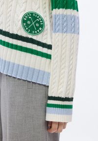 Εκρού πλεκτό πουλόβερ με πράσινες, μπλε και λευκές ρίγες· διαθέτει υφή πλέξης με κοτσίδες και στρογγυλή τσόχινη ετικέτα με λογότυπο στο μπροστινό μέρος.