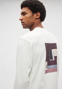 Balts, garām piedurknēm džemperis ar taisnstūra formas grafisko attēlu aizmugurē, kurā redzamas abstraktas formas un tonētas krāsas.