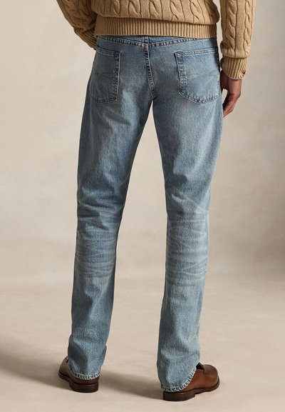 Bagfrafsyn af en person iført lyseblå jeans, en beige strikket sweater og brune lædersko, stående på en neutral baggrund.
