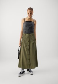 Crop top en denim avec des détails en clous, jupe maxi olive avec des boutons sur le devant, tissu texturé et sneakers noires.