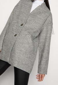 Grauer Tweedmantel mit lockerem Schnitt, langen Ärmeln und zwei Fronttaschen; verfügt über zwei dunkle Knöpfe und ein strukturiertes Muster.