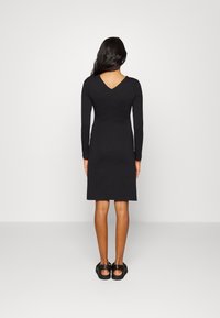 Vestido preto de mangas longas com uma silhueta ajustada e design nas costas em V, feito de um tecido suave. Combinado com sandálias pretas de bico aberto.
