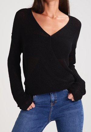 Femme portant un pull en maille noir à col en V avec manches longues, associé à un jean bleu, les mains dans les poches.