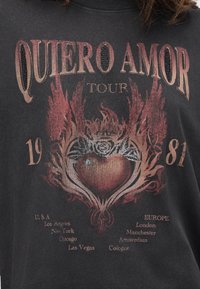 Μαύρο πουκάμισο με την επιγραφή "Quiero Amor Tour 1981," μια φλεγόμενη καρδιά με ένα στέμμα και τριαντάφυλλο, και ονόματα πόλεων από τις ΗΠΑ και την Ευρώπη από κάτω.