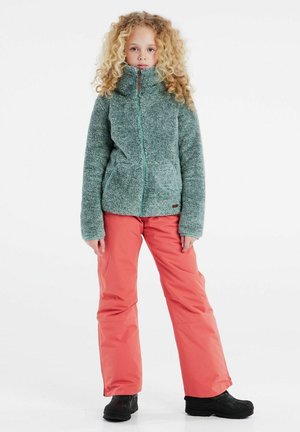 Giacca in fleece color teal con colletto alto e zip frontale, abbinata a pantaloni corallo. Stivali neri completano l'outfit da esterno.