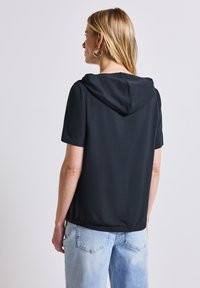 Top negro de manga corta con capucha, que presenta un dobladillo elástico. La tela parece suave, combinado con unos vaqueros azul claro.