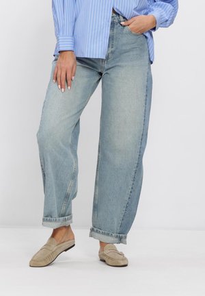 Vrouw die een lichtblauw gestreept overhemd draagt, losse lichtgewassen spijkerbroek met opgerolde zomen en beige instap loafers, poserend met één hand in de zak.