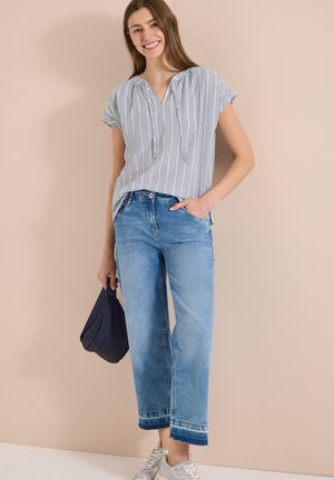 Junge Frau lächelt, trägt ein gestreiftes kurzärmeliges Oberteil, kurze blaue Jeans, weiße Sneakers, hält eine kleine dunkle Tasche in einer Hand.