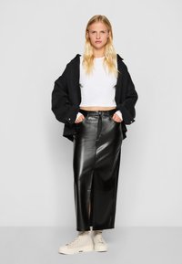 Zwarte leren maxi rok met een voorknipsel, gecombineerd met een witte cropped top en een oversized zwart jasje. Schoeisel is beige enkellaarsjes.
