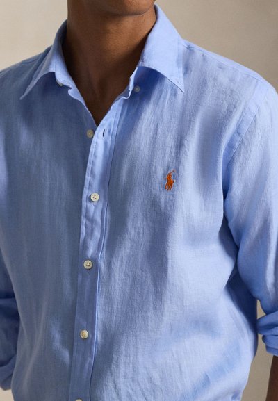 Μπλε λινό πουκάμισο με γιακά, λευκά κουμπιά και μικρό πορτοκαλί κεντημένο λογότυπο στο αριστερό στήθος. Ομαλή υφή.