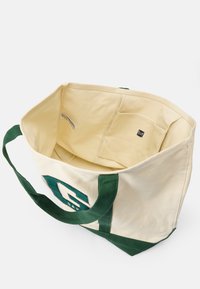 Saco de algodão em creme com detalhes em verde e logótipo. O interior possui um bolso e um forro suave. Alças duplas em tecido para transporte.