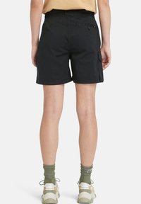 Shorts cargo neri con vestibilità dritta, dotati di tasca posteriore con bottone, passanti per cintura e una trama liscia. Indossati con calze verdi e sneakers.