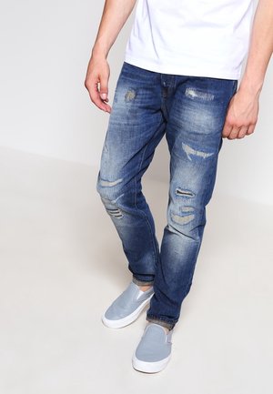 Hombre con vaqueros azules descoloridos y rotos, camiseta blanca y zapatos de lona gris claro, de pie sobre un suelo y fondo lisos y claros.