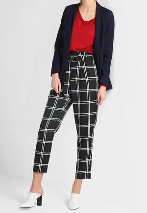 Femme portant un pantalon à carreaux noirs et blancs avec une ceinture, un haut rouge, un blazer bleu marine, et des mules blanches à talons, debout devant un fond blanc.