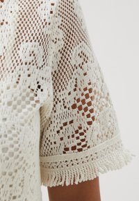 Haut en crochet blanc avec un design en maille ouverte, présentant des manches courtes et des accents de franges décoratives le long de l'ourlet.